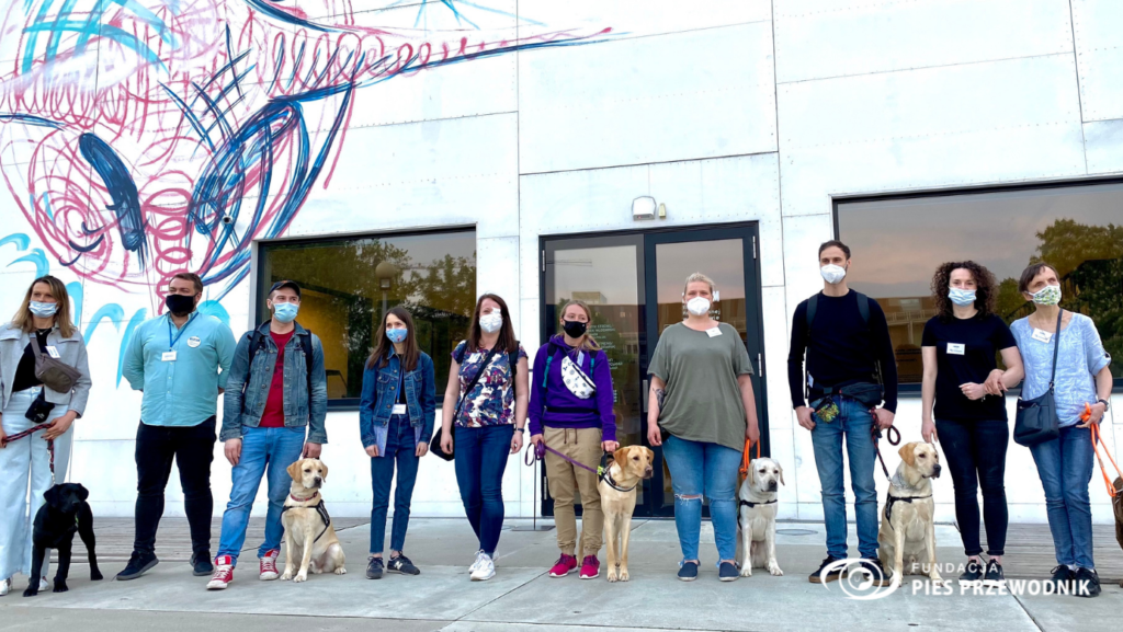 Przed budynkiem muzeum sztuki nowoczesnej stoi grupa 10 osób z 5 psami przewodnikami 